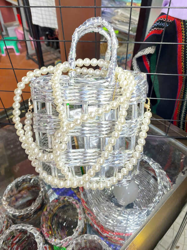 Một chiếc túi xách nhỏ được đan thủ công từ vỏ mì tôm có màu ánh kim bạc. Túi có quai xách ngắn làm từ cùng chất liệu và một quai đeo dài hơn kết từ những hạt ngọc trai nhân tạo màu trắng. Túi đang được trưng bày trên một bề mặt có nhiều sản phẩm tương tự khác.